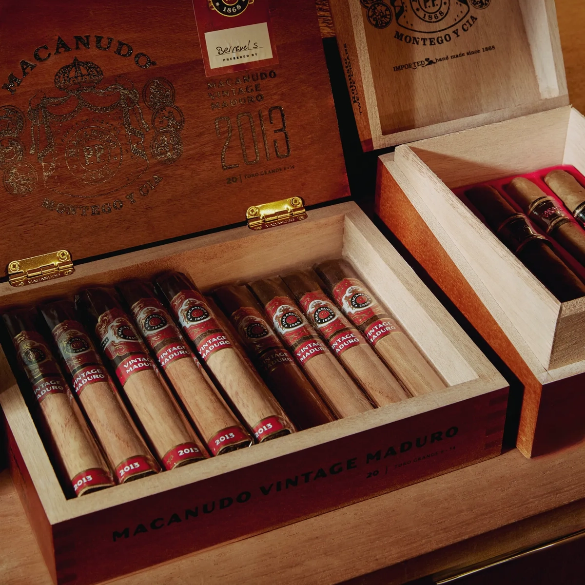 Open Macanudo cigar boxes on a shelf with vintage maduro sticks neatly lined, brass hinges and logos visible.