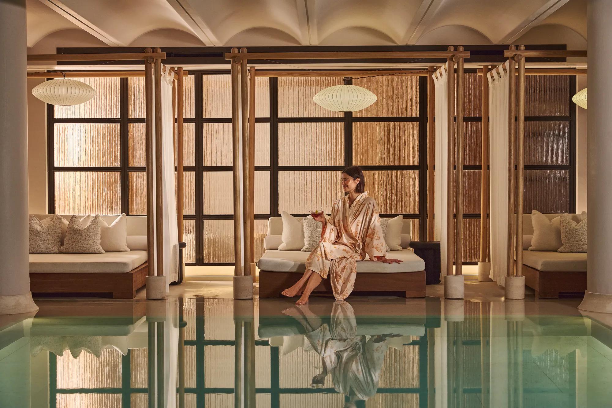 Person in a robe seated beside an indoor spa pool, with private cabanas and warm ambient lighting.