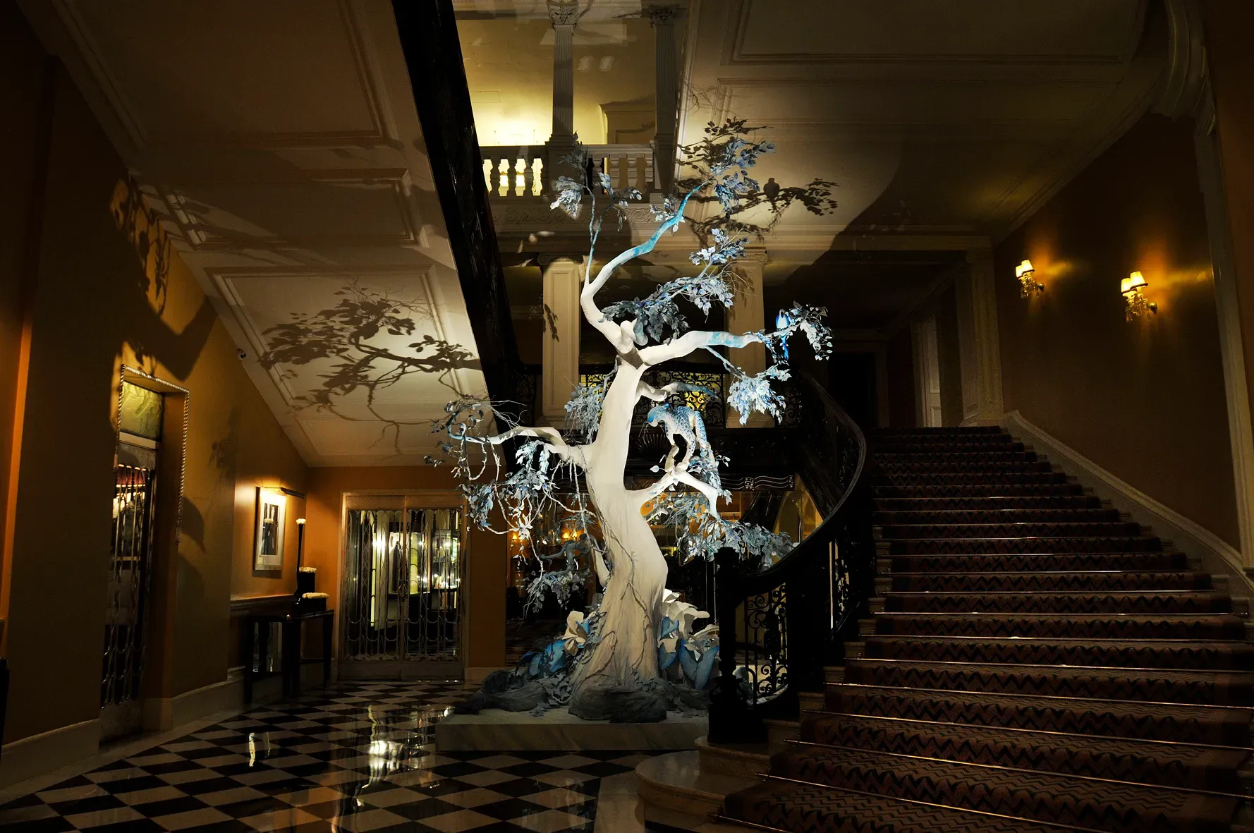 Grand hotel lobby with sweeping staircase and checkered floor, featuring sculptural white tree installation casting dramatic shadows under warm lighting.