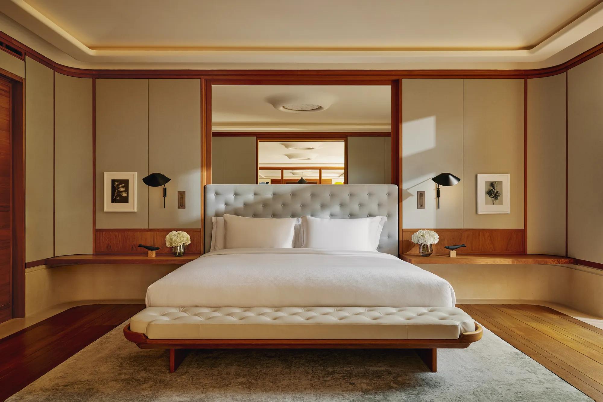 Bedroom in The Apartment featuring a king bed with a tall tufted headboard, flanked by black wall lights, with neutral tones and wood accents.