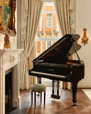 Elegant room with a black grand piano beside an ornate white fireplace, framed by tall draped curtains and a window overlooking red brick architecture.