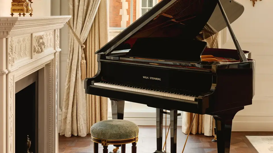 Elegant room with a black grand piano beside an ornate white fireplace, framed by tall draped curtains and a window overlooking red brick architecture.