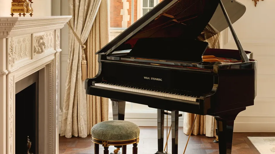 Elegant room with a black grand piano beside an ornate white fireplace, framed by tall draped curtains and a window overlooking red brick architecture.