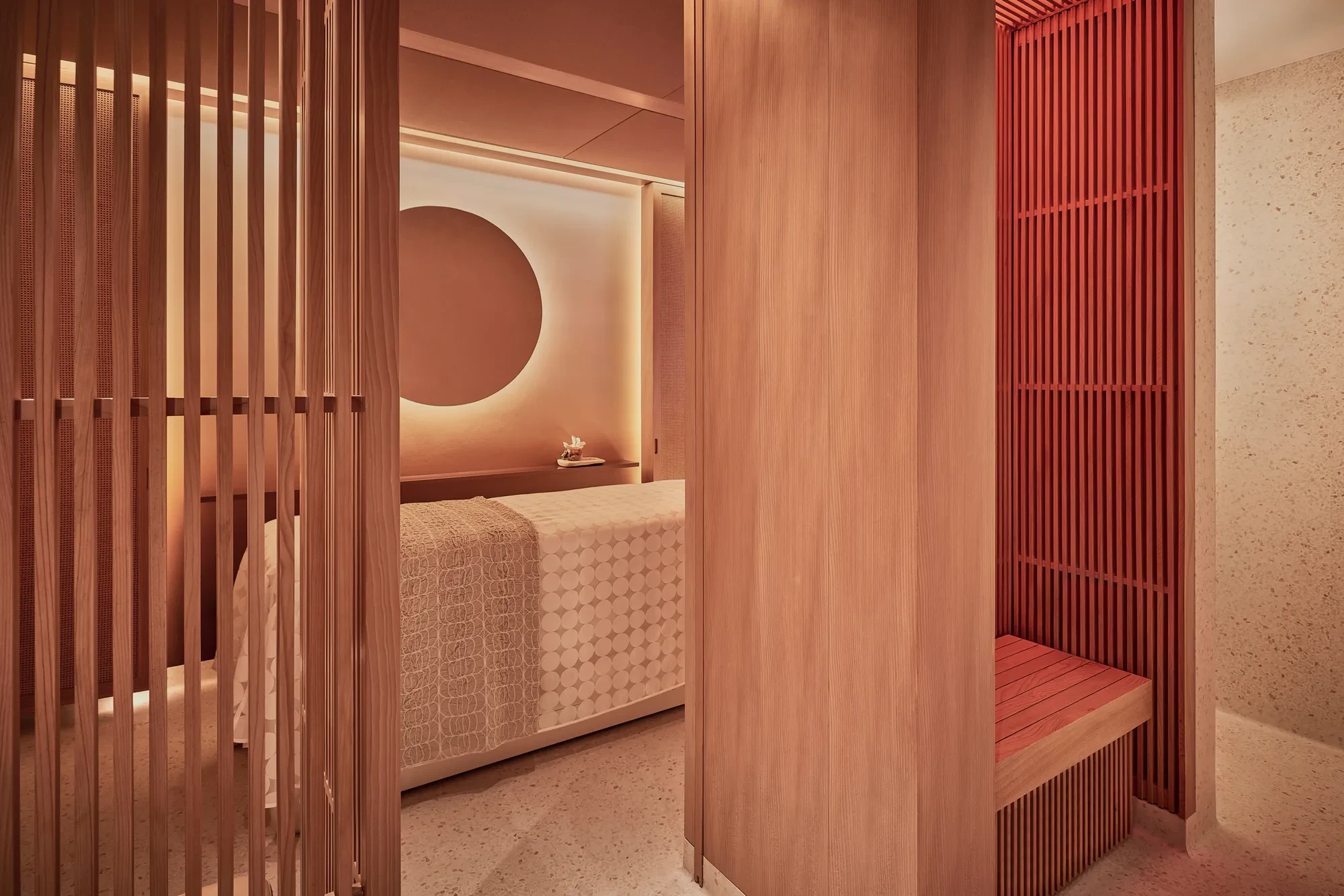 Warm-toned spa treatment room with massage bed, wooden slatted screens and softly lit feature wall.