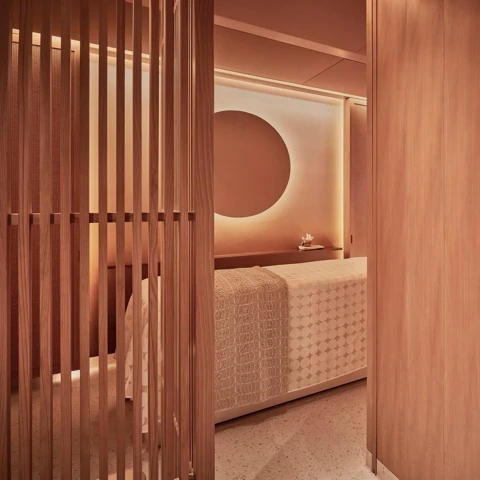 Warm-toned spa treatment room with massage bed, wooden slatted screens and softly lit feature wall.