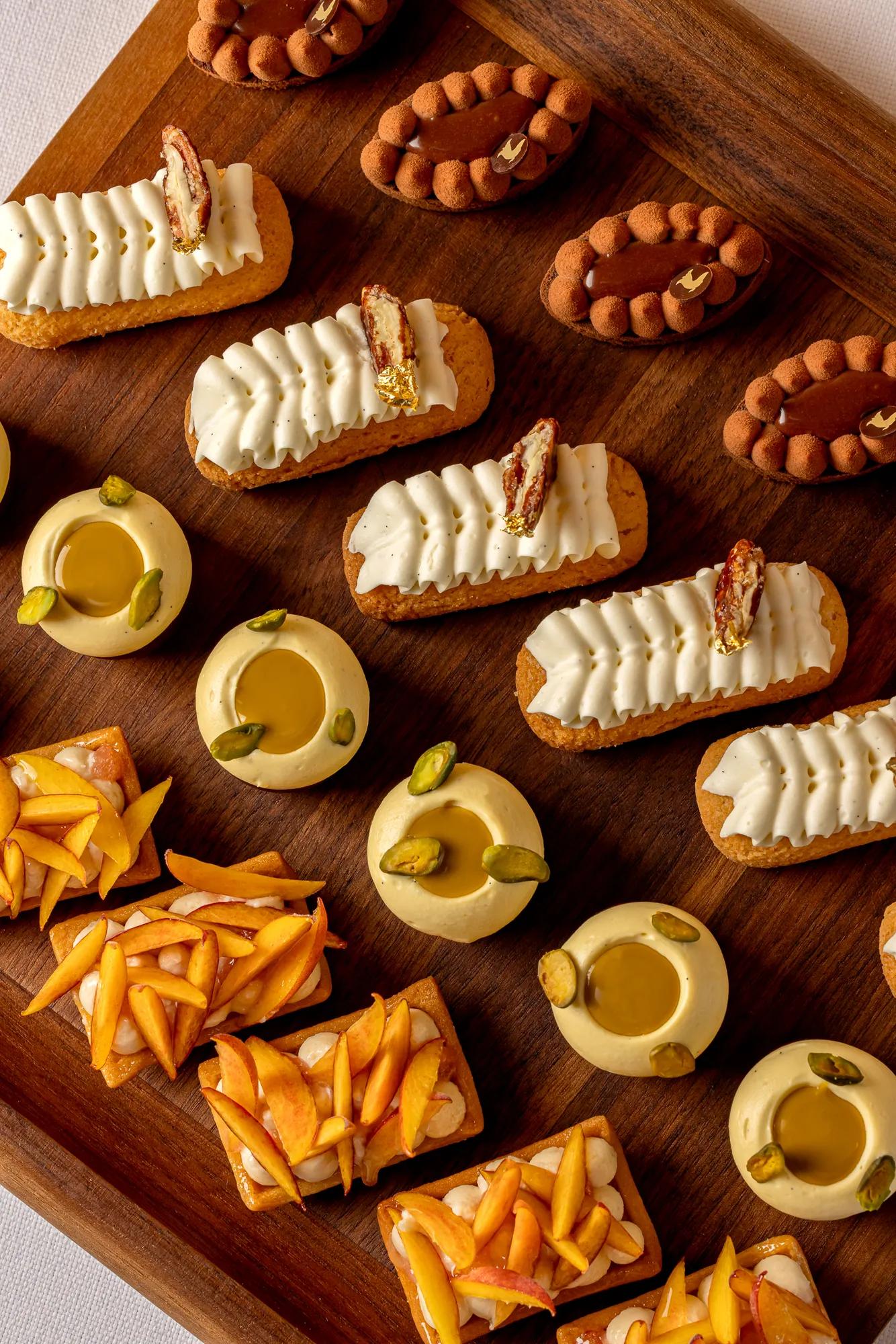 Assorted bite-sized pastries arranged on a wooden board, including eclairs, fruit tarts, and round mousse cakes with pistachio.