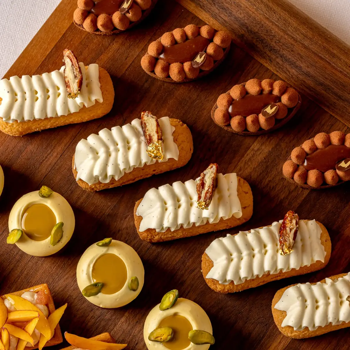 Assorted bite-sized pastries arranged on a wooden board, including eclairs, fruit tarts, and round mousse cakes with pistachio.