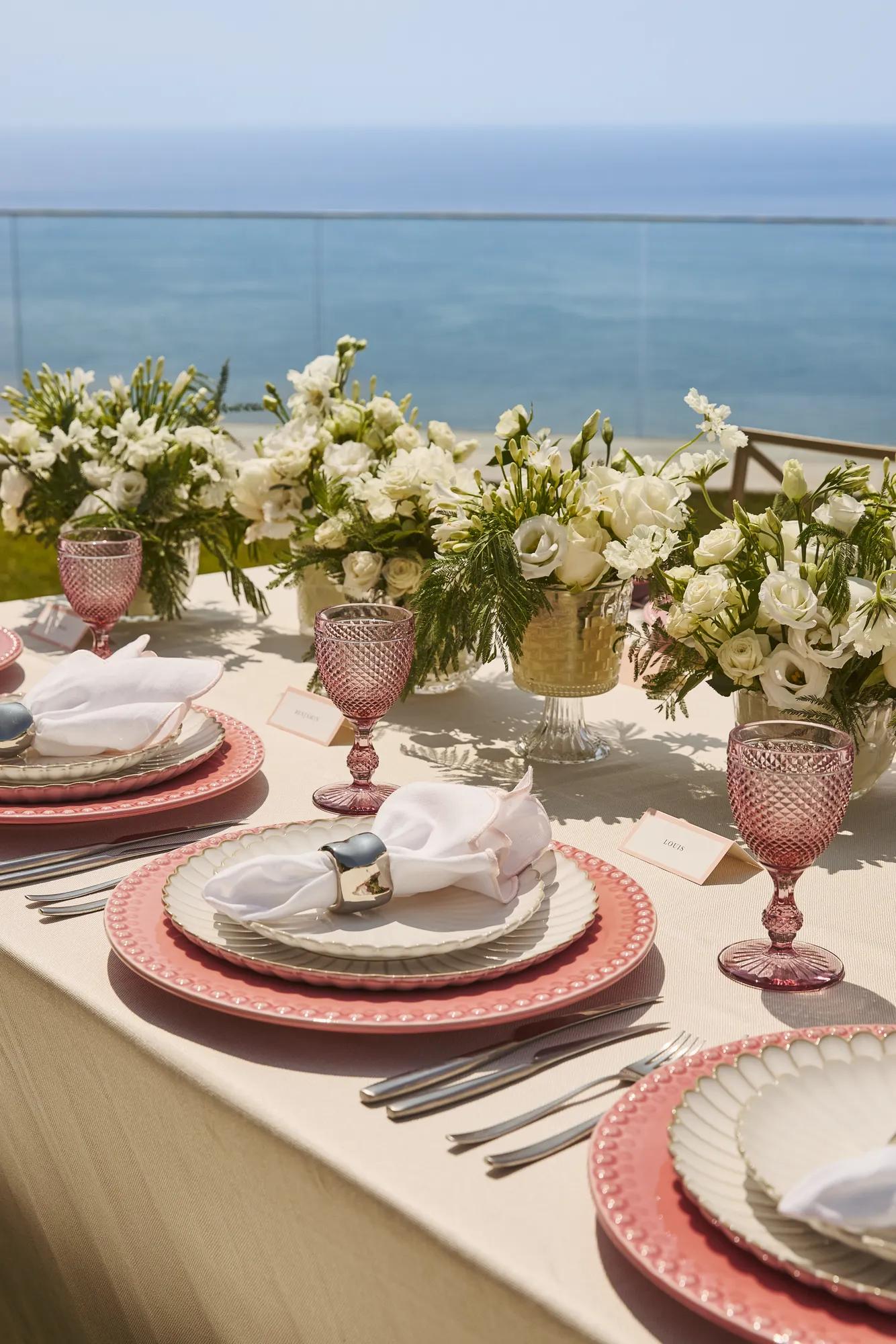 Elegantly set outdoor table with pink glassware, layered plates and white floral arrangements, overlooking calm sea under clear sky.