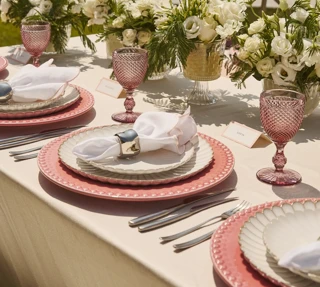 Elegantly set outdoor table with pink glassware, layered plates and white floral arrangements, overlooking calm sea under clear sky.