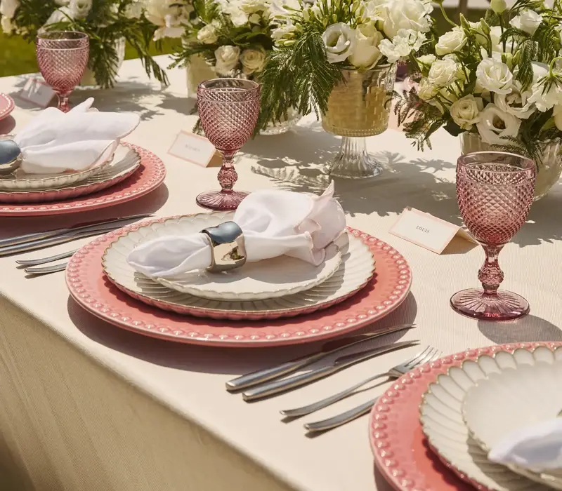 Elegantly set outdoor table with pink glassware, layered plates and white floral arrangements, overlooking calm sea under clear sky.