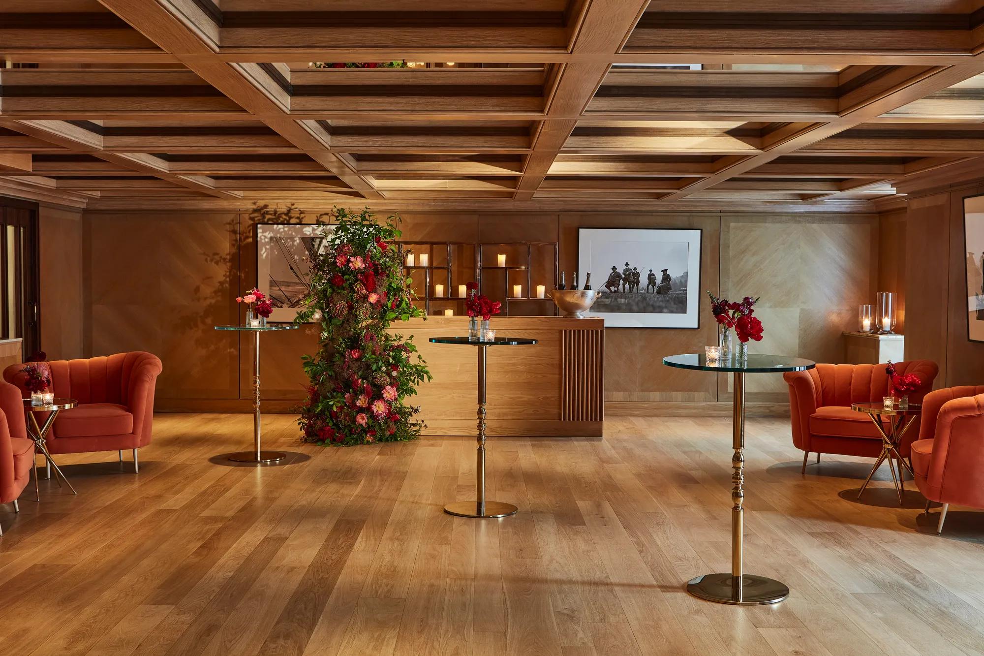 Warm event lounge with glass-topped cocktail tables, orange armchairs, and a floral display beside a candle-lit bar.