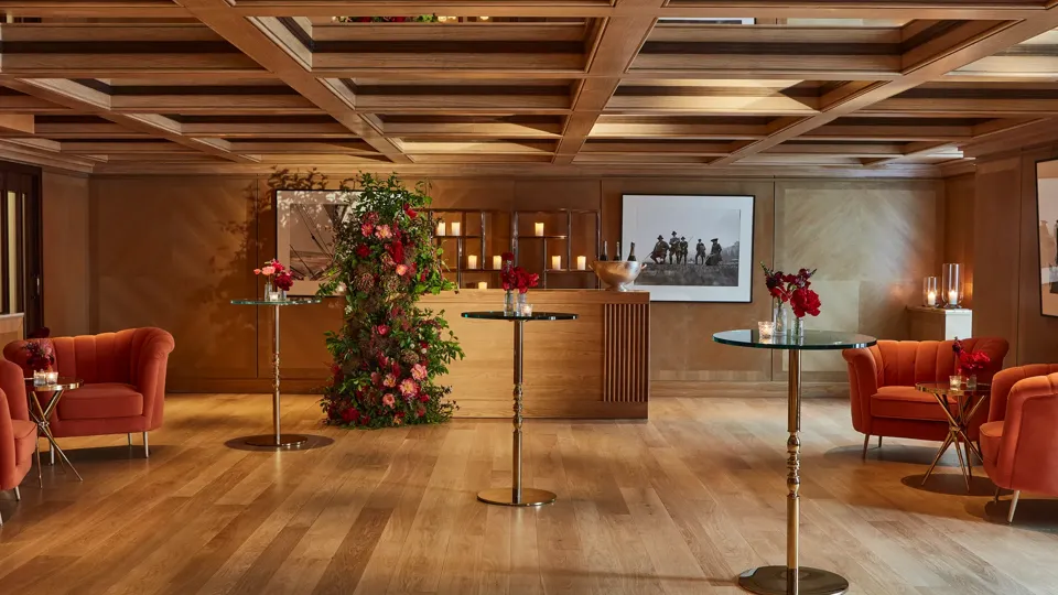 Warm event lounge with glass-topped cocktail tables, orange armchairs, and a floral display beside a candle-lit bar.