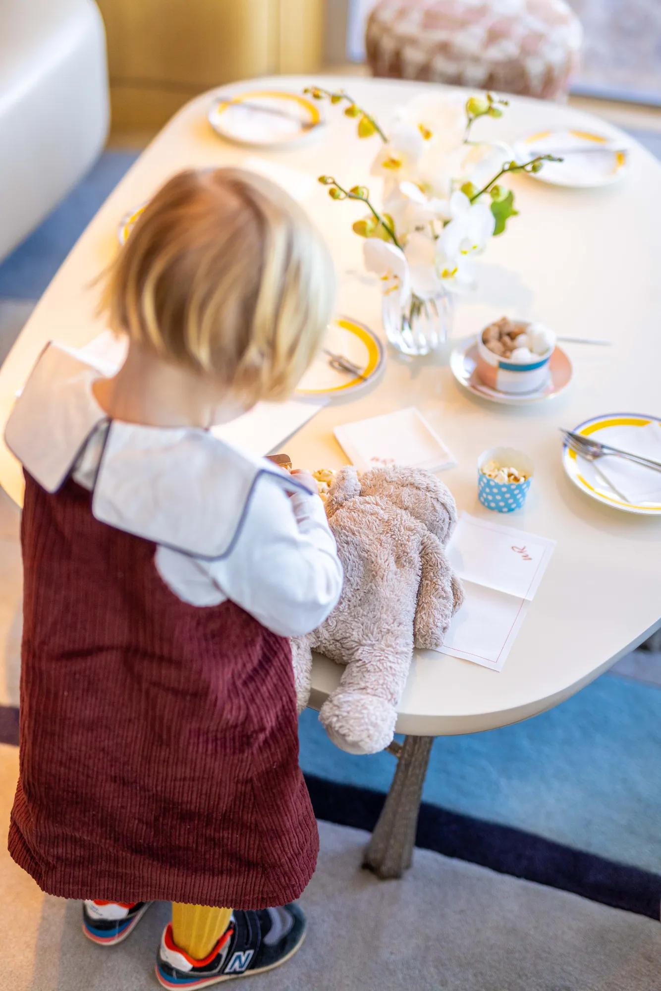 Un jeune enfant avec une peluche se tient devant une table joliment dressée, ornée de fleurs et de vaisselle.
