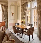 Elegant seating area with a wooden dining table set for four beside tall windows with patterned drapes, gold-trimmed wall panels, and a vase of pink flowers as the centerpiece.