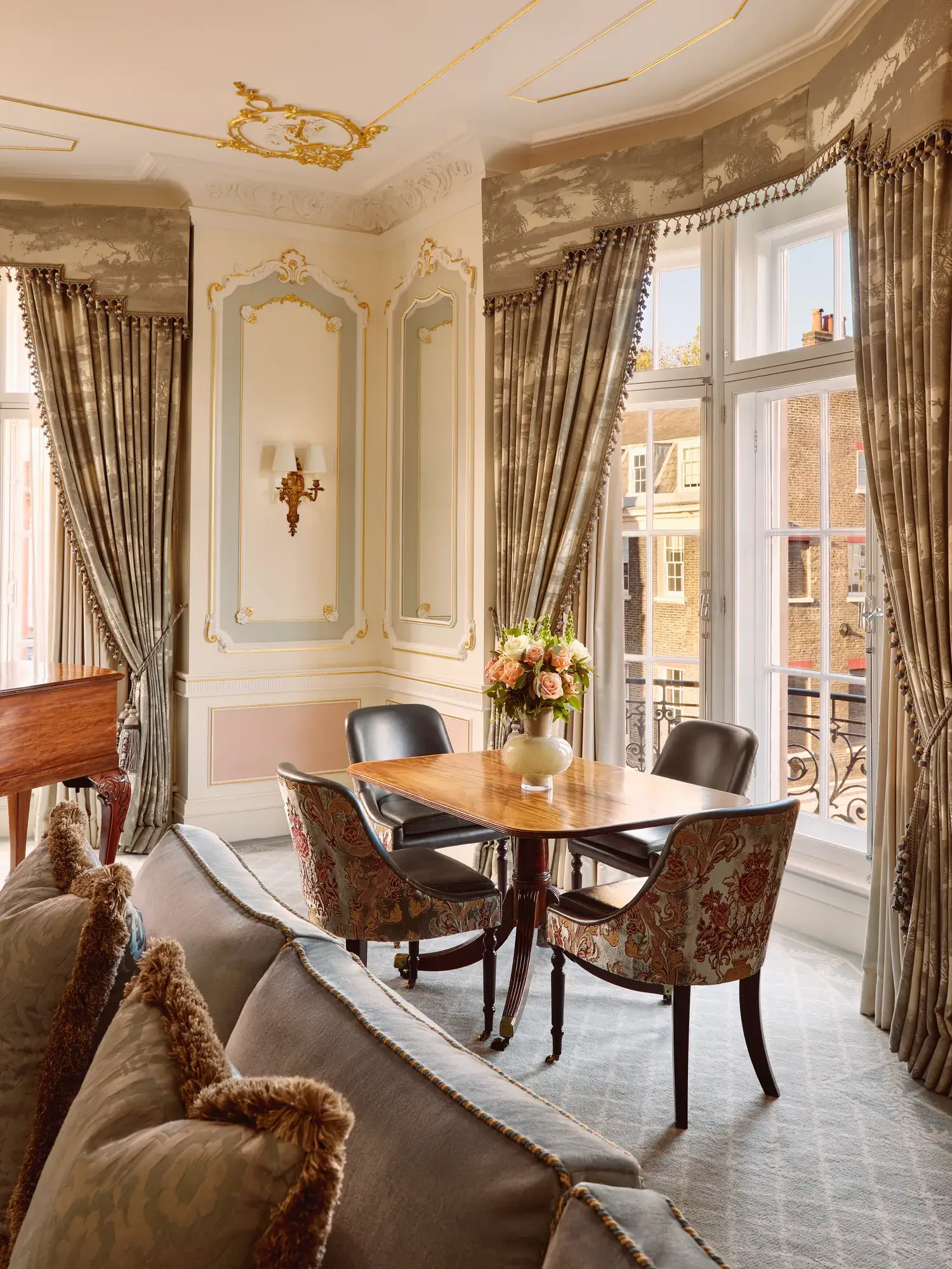 Elegant seating area with a wooden dining table set for four beside tall windows with patterned drapes, gold-trimmed wall panels, and a vase of pink flowers as the centerpiece.