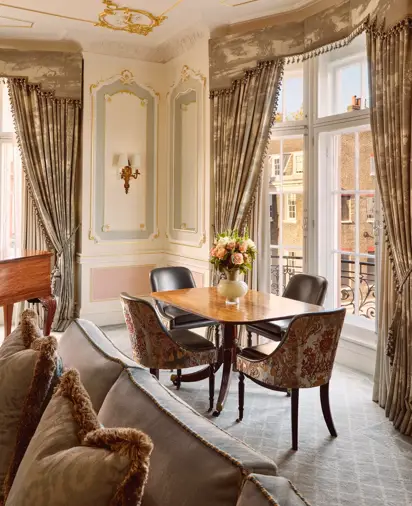 Elegant seating area with a wooden dining table set for four beside tall windows with patterned drapes, gold-trimmed wall panels, and a vase of pink flowers as the centerpiece.