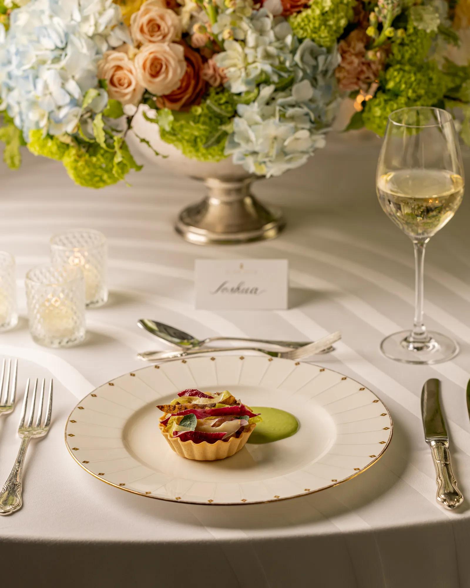 Individual tartlet with colourful vegetable garnish and green purée, served on a formally set table beneath a floral centrepiece.