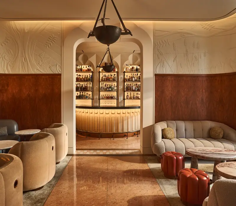 Elegant lounge with curved seating and marble tables, opening onto a softly lit bar framed by an arched doorway.