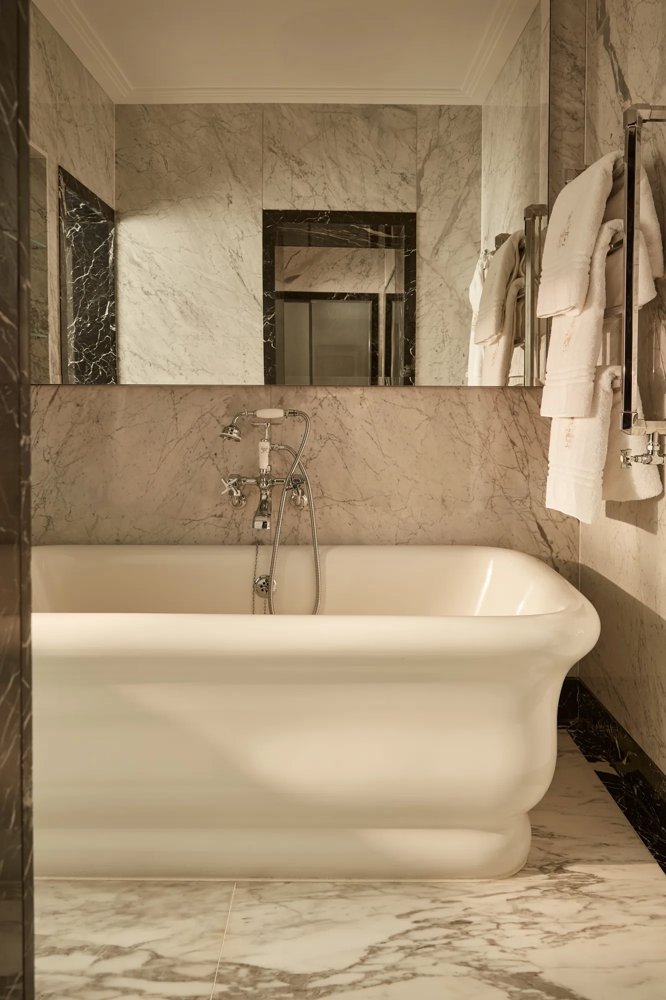Elegant marble bathroom with a freestanding white bathtub, wall-mounted taps, and plush towels neatly hung on a chrome rail under soft ambient lighting.