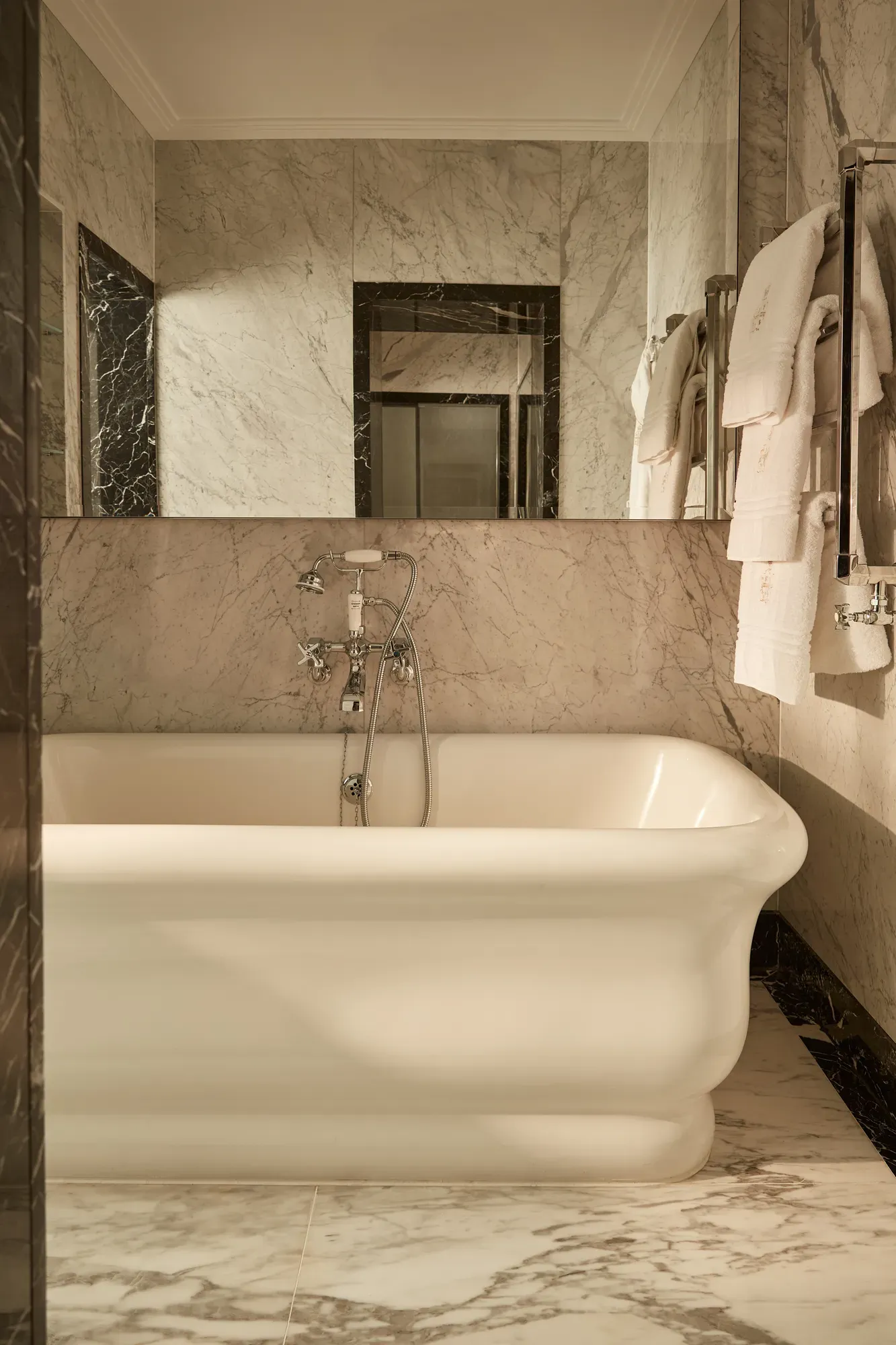 Elegant marble bathroom with a freestanding white bathtub, wall-mounted taps, and plush towels neatly hung on a chrome rail under soft ambient lighting.