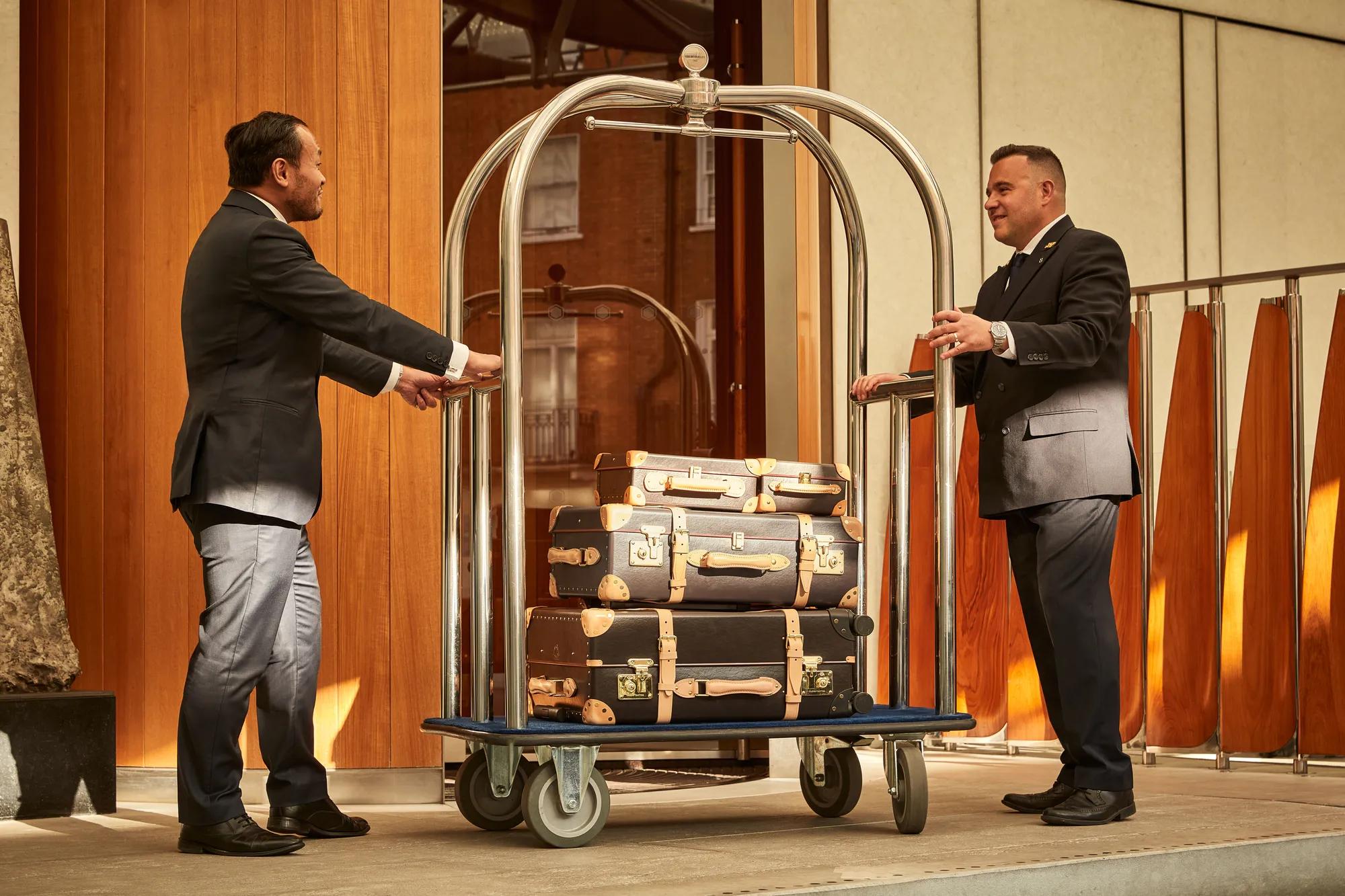 Hotel staff standing by a luggage trolley stacked with vintage-style suitcases at the entrance of a luxury hotel.