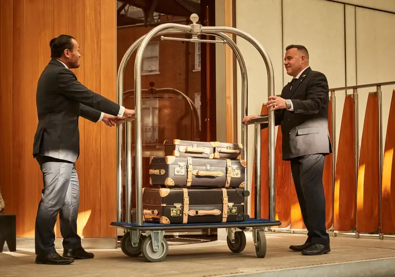 Hotel staff standing by a luggage trolley stacked with vintage-style suitcases at the entrance of a luxury hotel.
