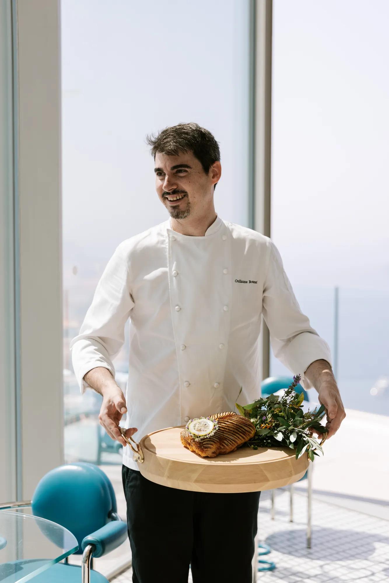 Un chef en veste blanche présente un plat de poisson dressé sur une planche en bois dans un restaurant baigné de lumière.