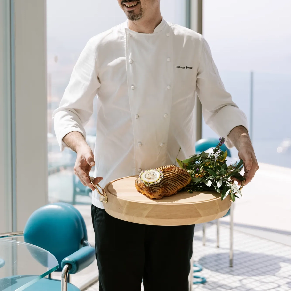 Un chef en veste blanche présente un plat de poisson dressé sur une planche en bois dans un restaurant baigné de lumière.