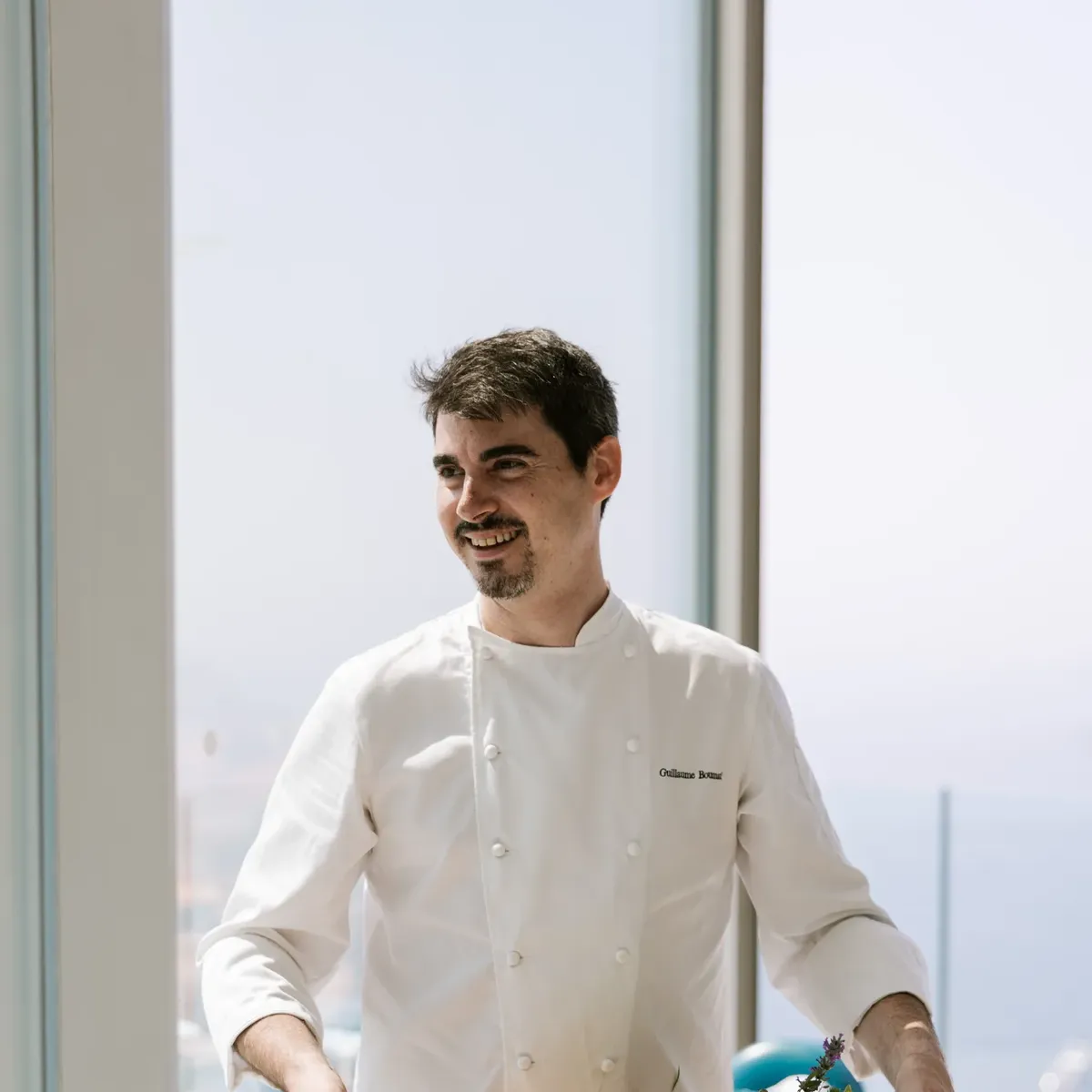 Un chef en veste blanche présente un plat de poisson dressé sur une planche en bois dans un restaurant baigné de lumière.
