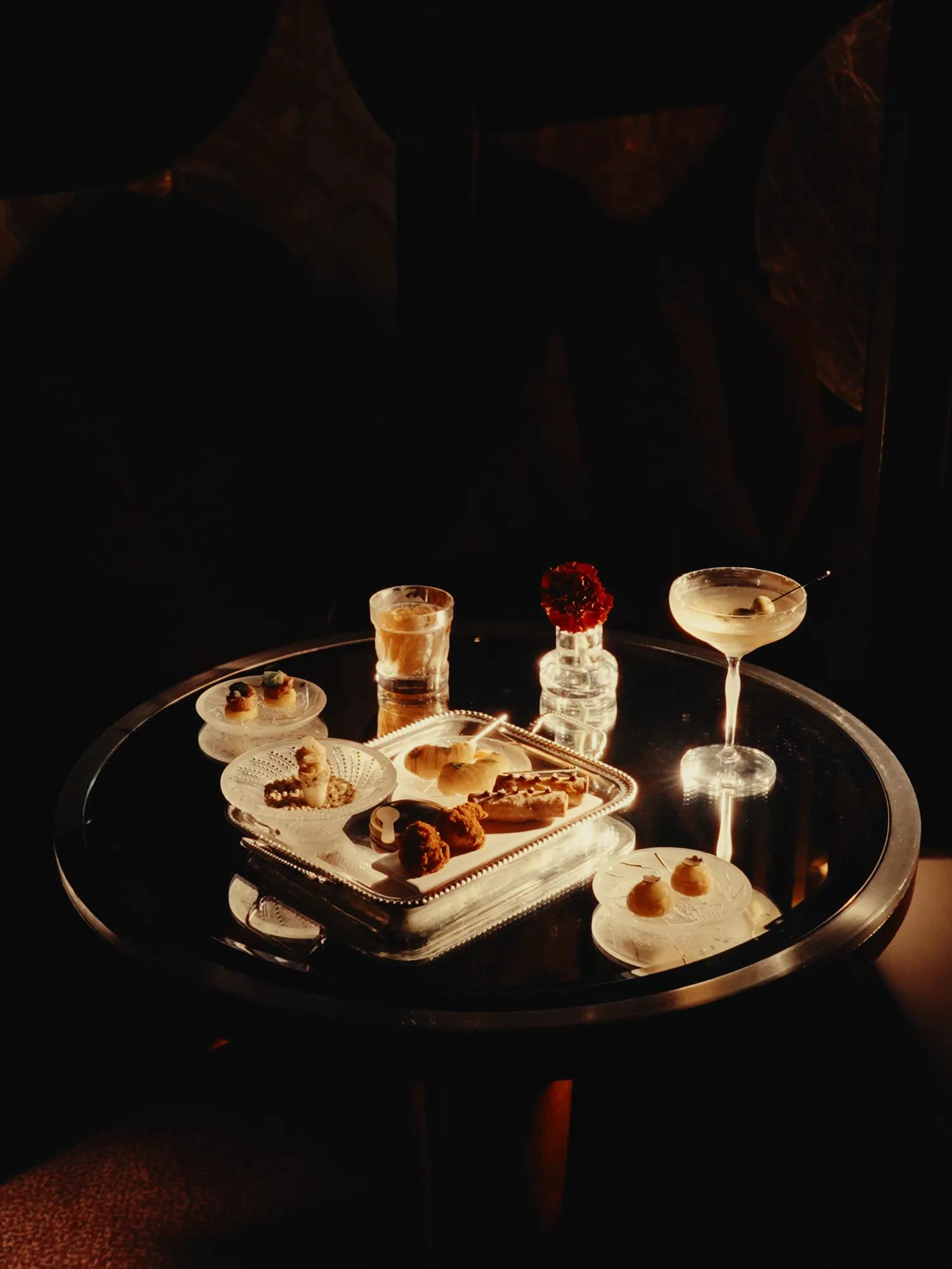 A selection of cocktails and refined savoury bites arranged on a softly lit table, creating an intimate atmosphere for evening drinks and sharing plates.