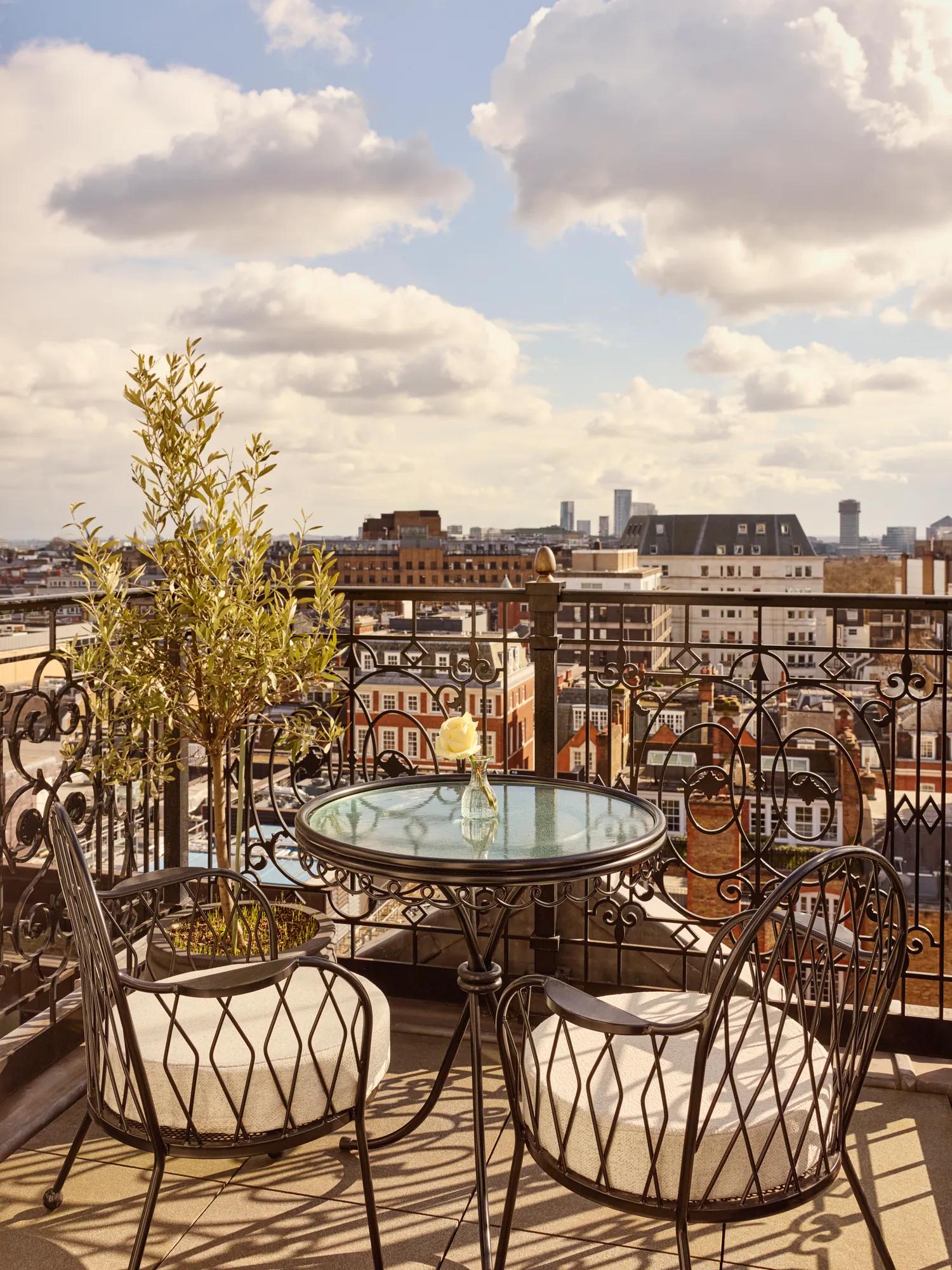 Small terrace with two cushioned chairs and a round glass table overlooking city rooftops under a cloudy sky.