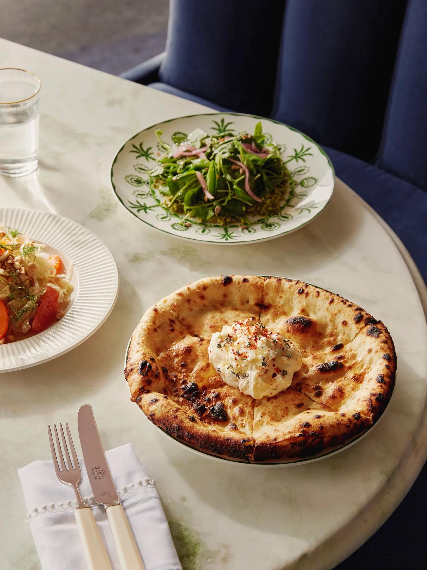 Charred wood-fired flatbread topped with whipped ricotta; light salads blurred in the background.