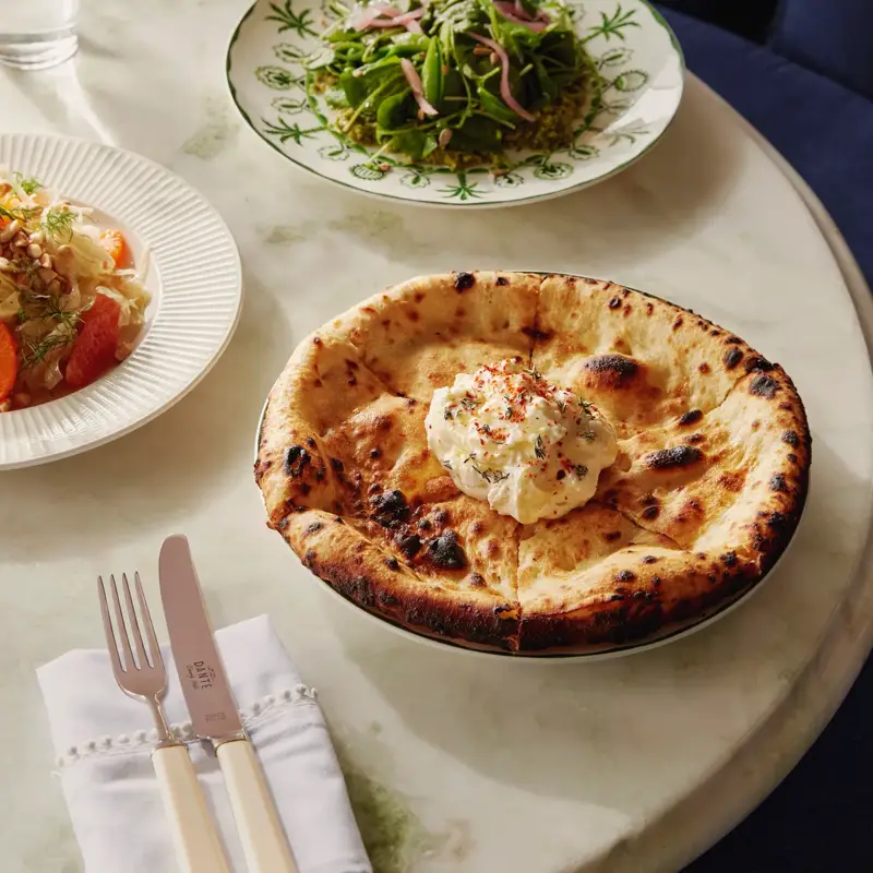Charred wood-fired flatbread topped with whipped ricotta; light salads blurred in the background.