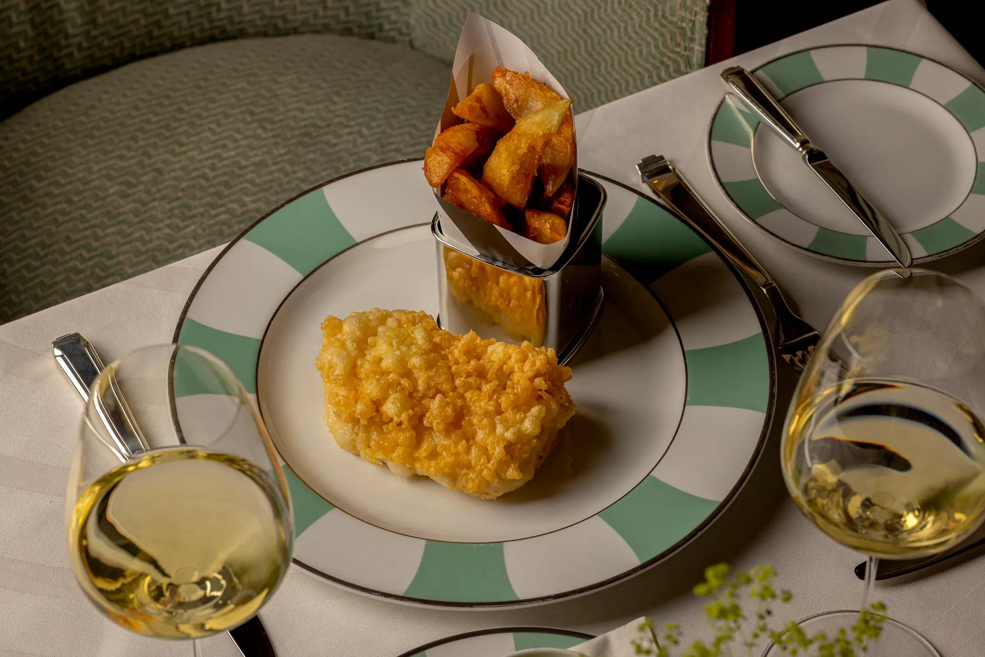 Breaded fish fillet with chunky chips on a patterned plate, accompanied by glasses of white wine.