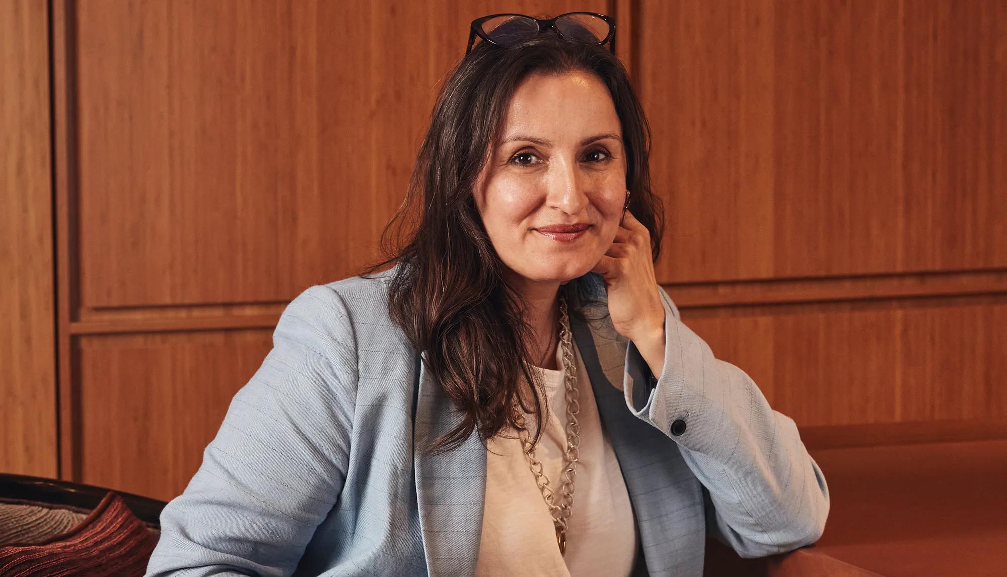 Woman in light blue blazer seated against warm wood panel wall, resting chin on hand and smiling.