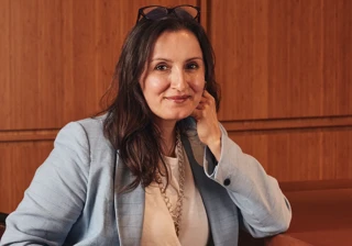 Woman in light blue blazer seated against warm wood panel wall, resting chin on hand and smiling.
