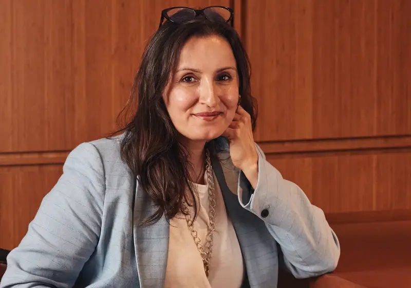 Woman in light blue blazer seated against warm wood panel wall, resting chin on hand and smiling.