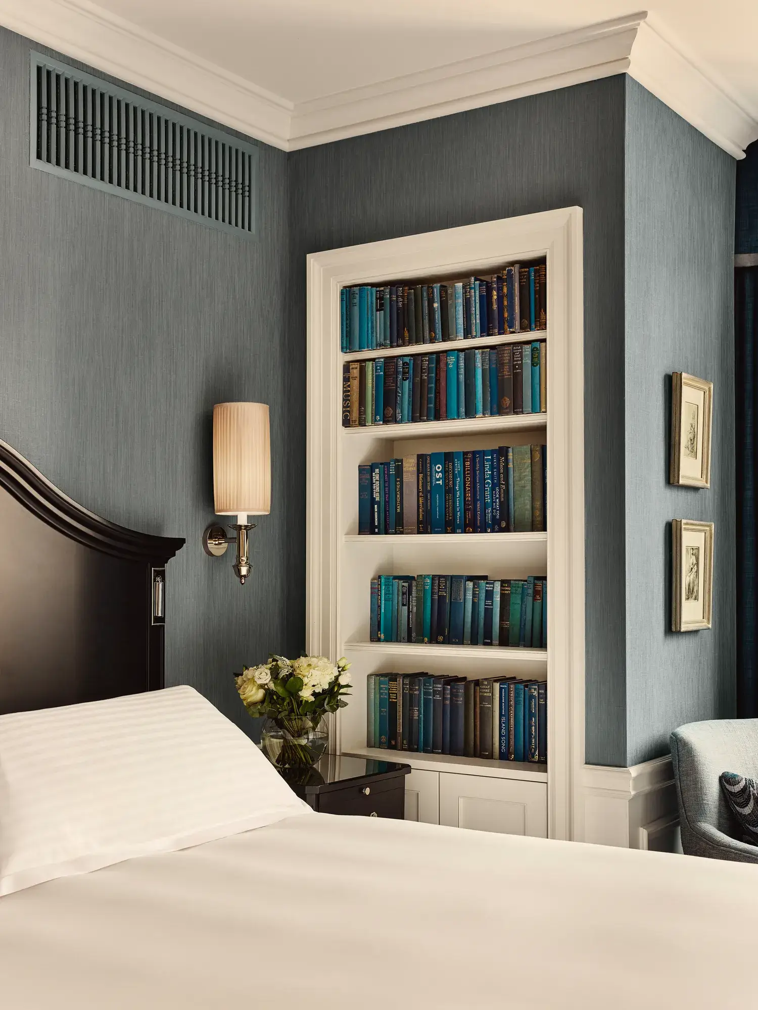 Bedroom corner with dark wood headboard, built-in bookshelf of blue books, wall sconce, and small bedside flowers.
