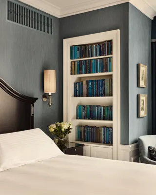 Bedroom corner with dark wood headboard, built-in bookshelf of blue books, wall sconce, and small bedside flowers.