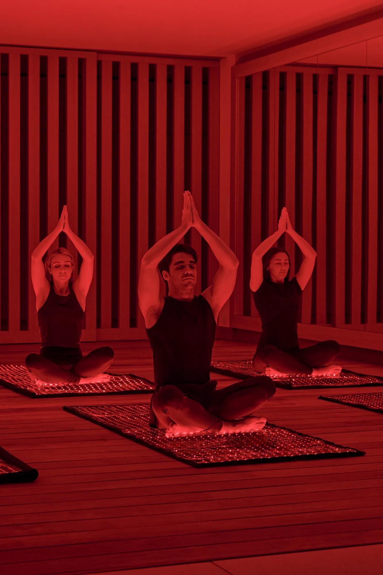 Three guests seated on mats in red-lit studio, performing seated yoga pose with hands pressed together overhead.