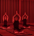 Three guests seated on mats in red-lit studio, performing seated yoga pose with hands pressed together overhead.