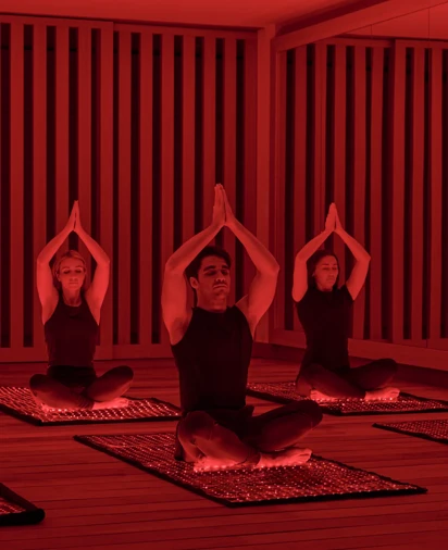 Three guests seated on mats in red-lit studio, performing seated yoga pose with hands pressed together overhead.