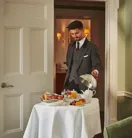 A suited server presents an elegant in-room breakfast on a white-clothed trolley, with pastries, fruit and tea arranged neatly in a softly lit suite.