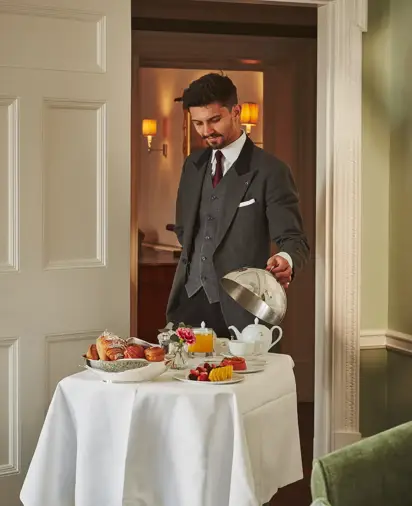 A suited server presents an elegant in-room breakfast on a white-clothed trolley, with pastries, fruit and tea arranged neatly in a softly lit suite.