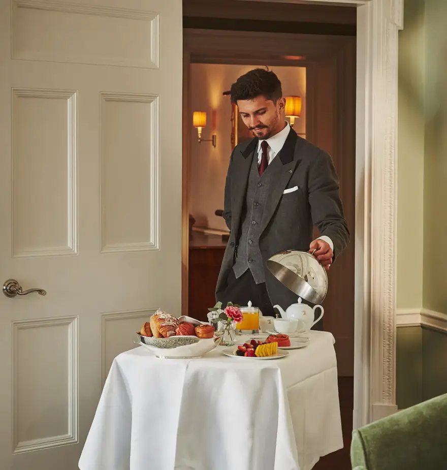 A suited server presents an elegant in-room breakfast on a white-clothed trolley, with pastries, fruit and tea arranged neatly in a softly lit suite.