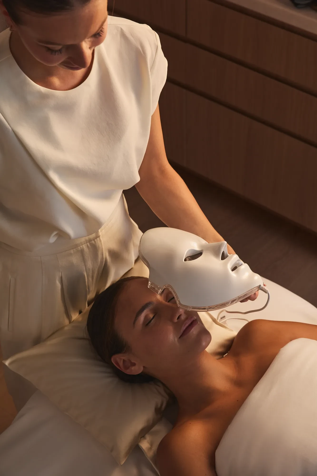 Spa therapist holding a light therapy mask above a guest during a facial treatment in a serene room.