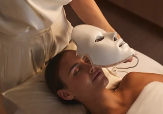 Spa therapist holding a light therapy mask above a guest during a facial treatment in a serene room.