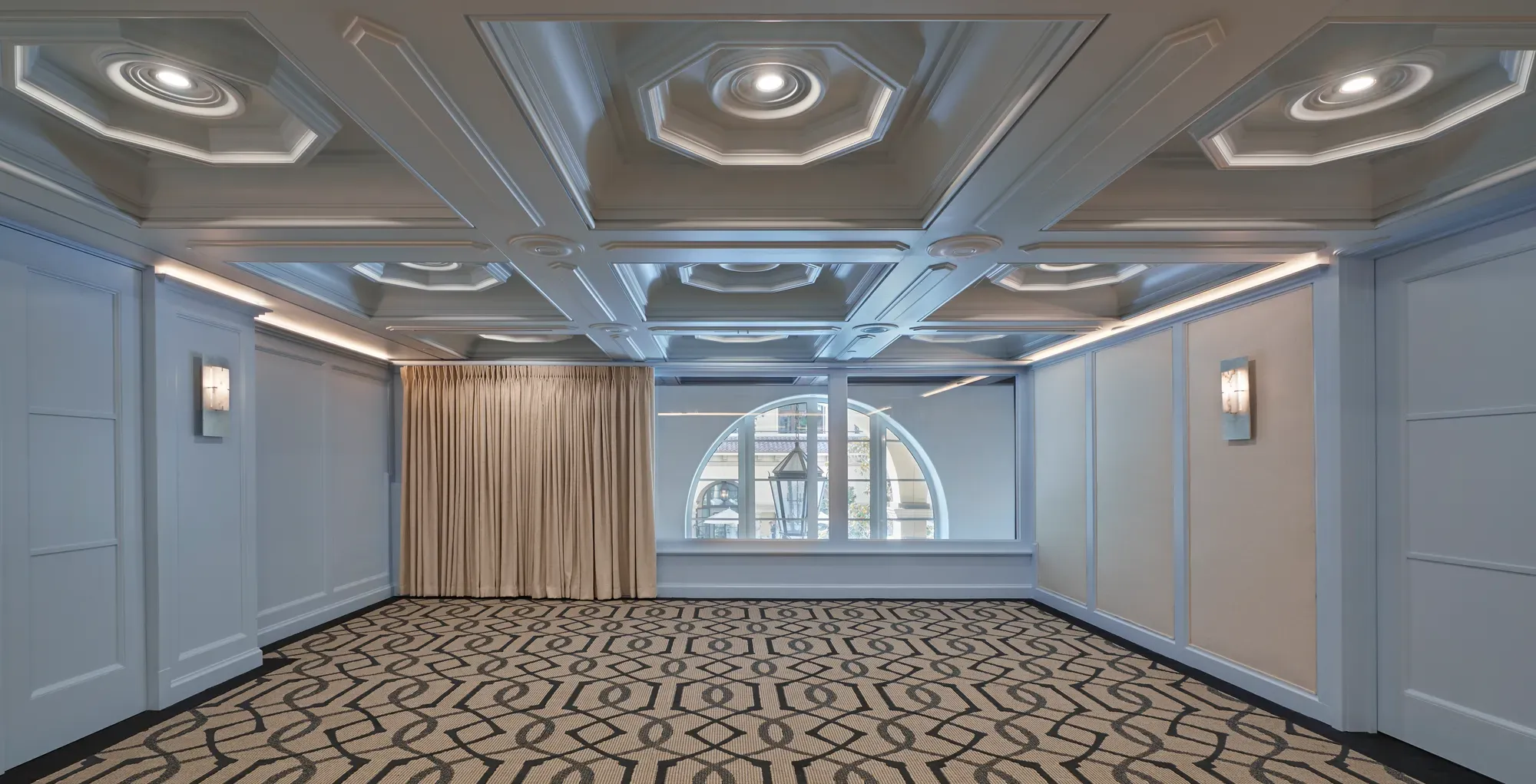 Empty Beverly room with patterned carpet, pale blue paneled walls, coffered ceiling, and arched window with drapes.