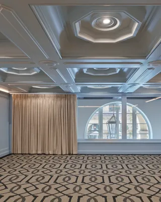 Empty Beverly room with patterned carpet, pale blue paneled walls, coffered ceiling, and arched window with drapes.