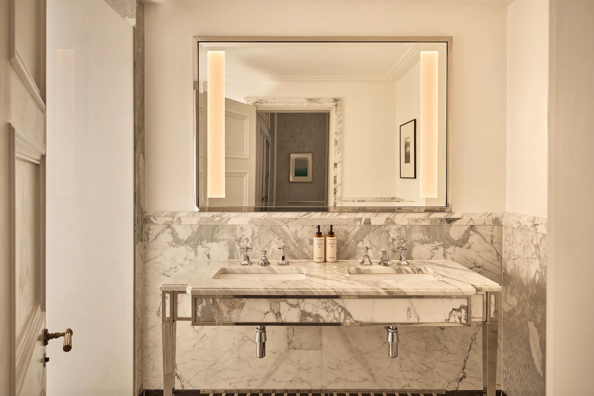 Marble double vanity with twin sinks, illuminated mirror and stone-clad walls, viewed through an open doorway into the bathroom.